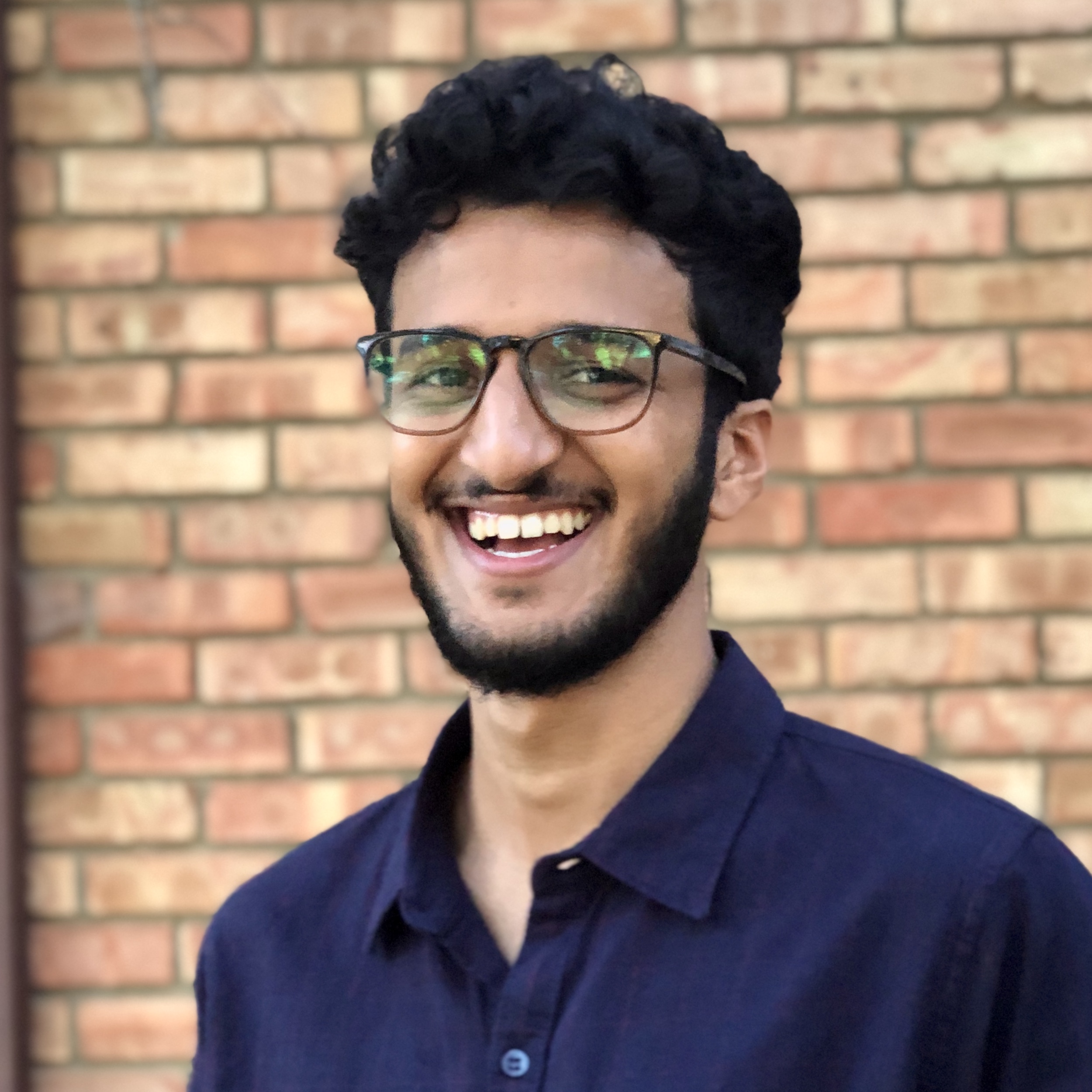 Photo of Hrishi. He is wearing a black check shirt and is smiling as he looks at the camera. He is standing in front of a brick wall